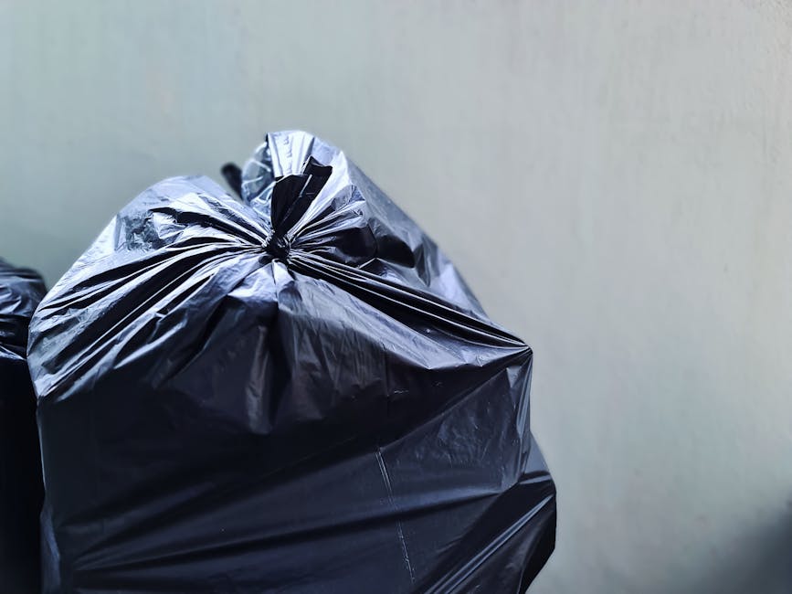 Close-up view of multiple large black plastic rubbish bags, tightly sealed with the tops gathered and knotted, stacked together against a plain, light-colored wall. The bags appear to be made from dur