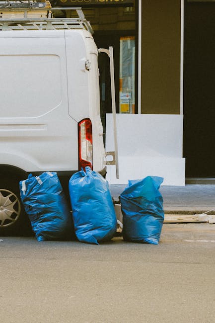 Close-up view of multiple large black plastic rubbish bags, tightly sealed with the tops gathered and knotted, stacked together against a plain, light-colored wall. The bags appear to be made from dur