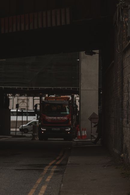 Underneath a concrete bridge supported by large, rectangular pillar columns with visible vertical grooves, a pile of dark, weathered wooden logs and branches is situated on the ground, partially surro